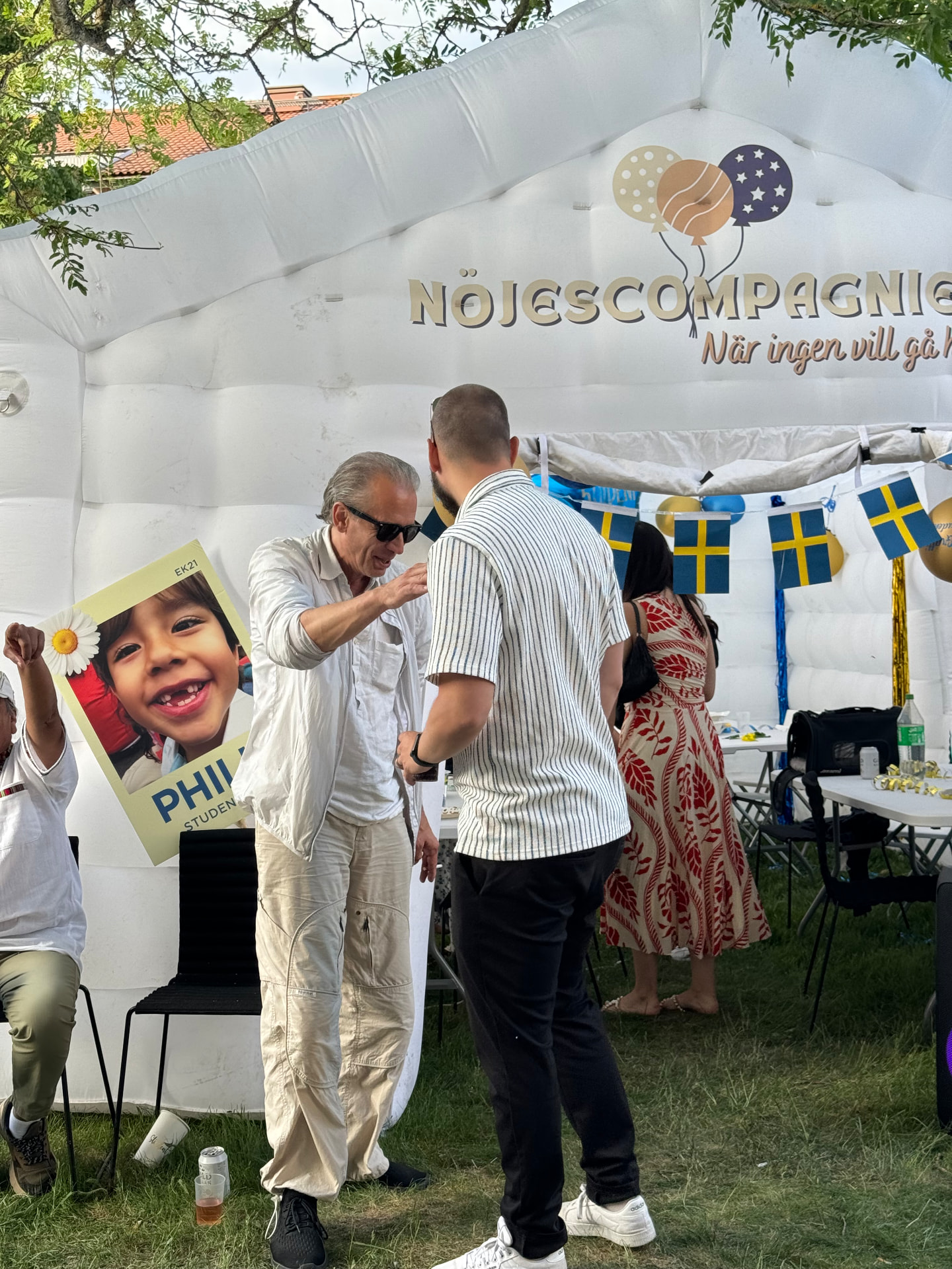 Gäster vid ett studentfirande står vid ingången till det vita uppblåsbara partytältet med svenska flaggor och ballonger