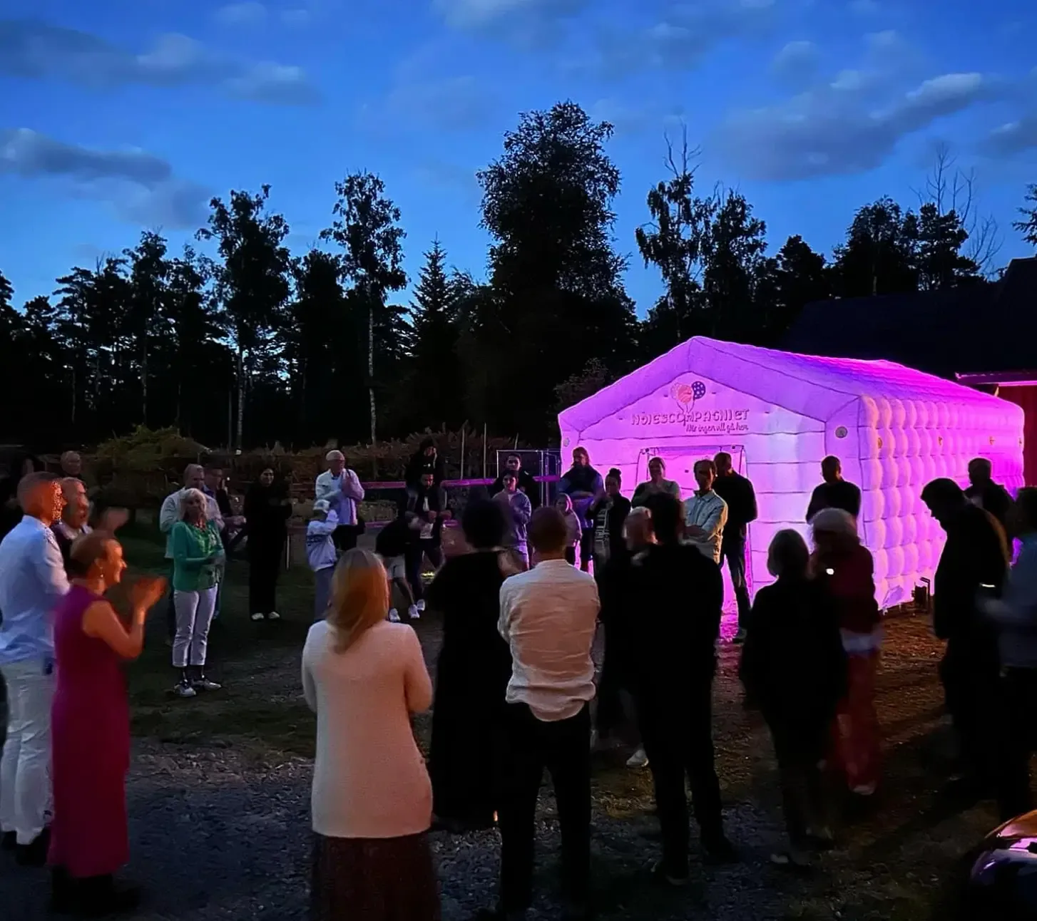 Vitt uppblåsbart partytält med lila belysning och festgäster samlade utanför i skymningen